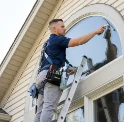 Professional window cleaner on a ladder using a squeegee to clean an exterior arched window, Chandler, AZ.